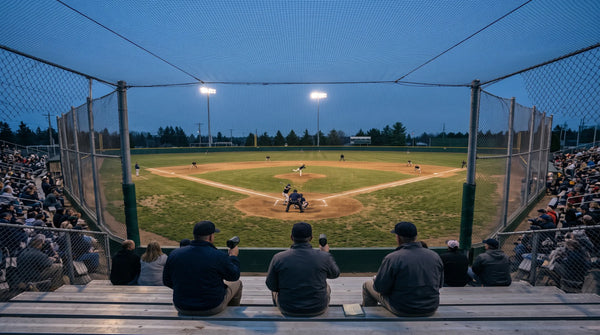 A baseball scout’s hands hold a radar gun and handwritten scouting notebook in the foreground as a young pitcher delivers to a batter during an evening game on a high school or college field.