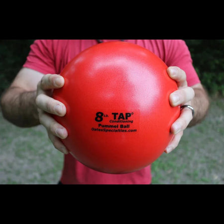 An athlete grips a red 8lb pummel ball with two hands showing the size and the textured finish.