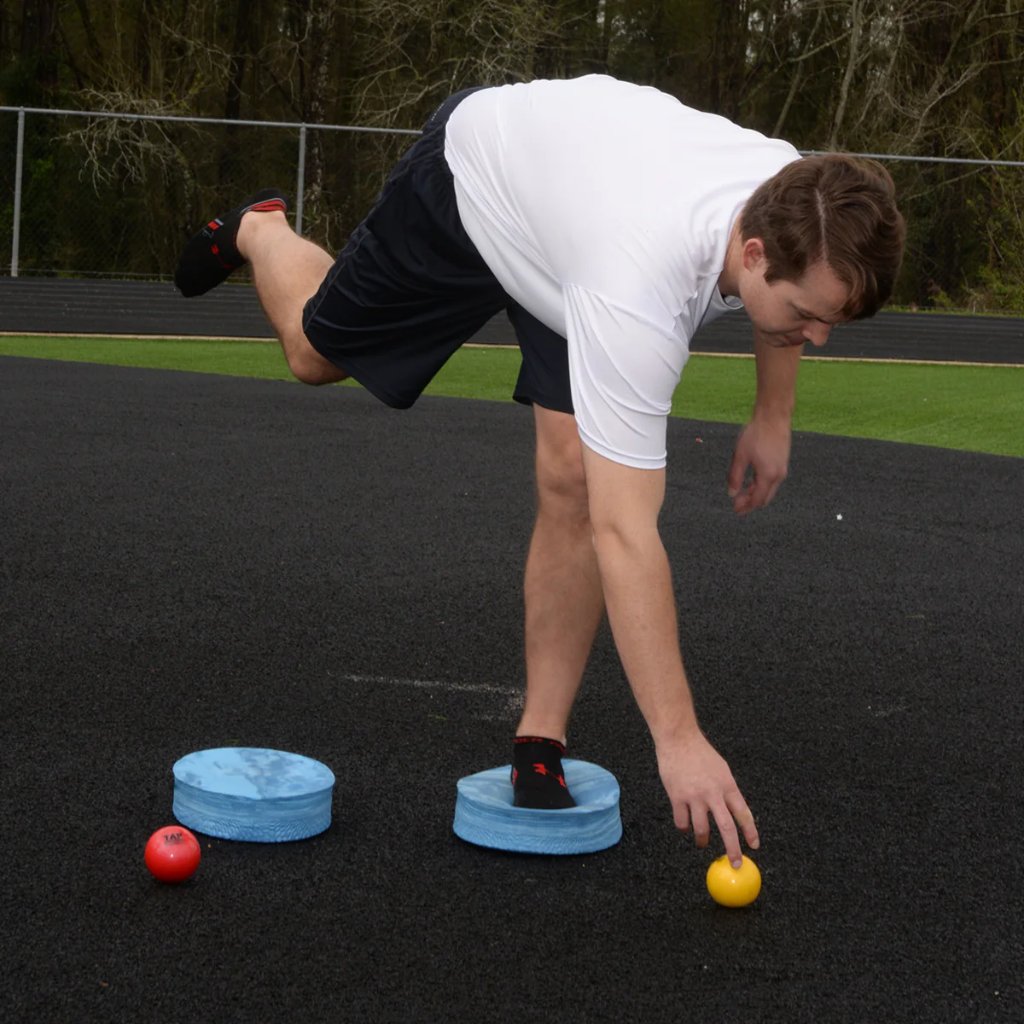 Athlete maintaining ankle stability on an Oval Balance Pad while reaching for a yellow ball, demonstrating balance, coordination, and controlled movement during training.