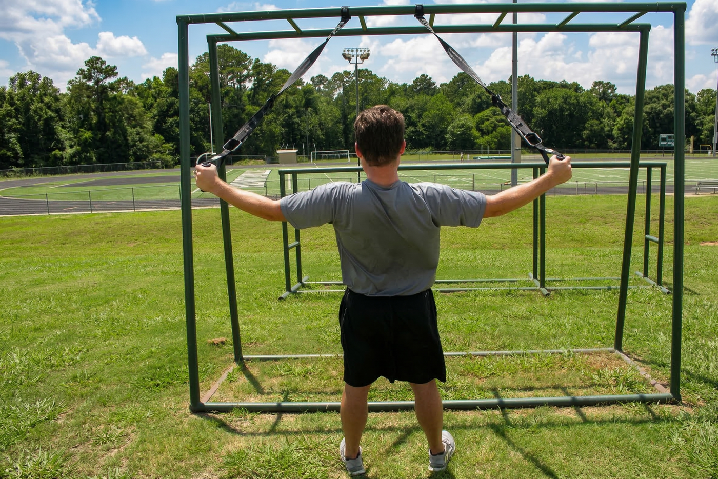 The man grips the rings attached to black suspension trainer straps, which hang from a metal exercise frame. He stands with his back to the viewer and looks straight ahead as he holds his arms out in a "T" pose.