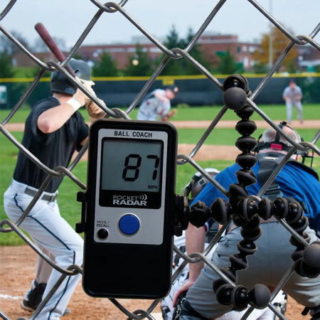 Ball Coach™ radar gun secured to a chain-link fence, actively tracking pitch speeds during a live baseball game—hands-free velocity data for in-game analysis.