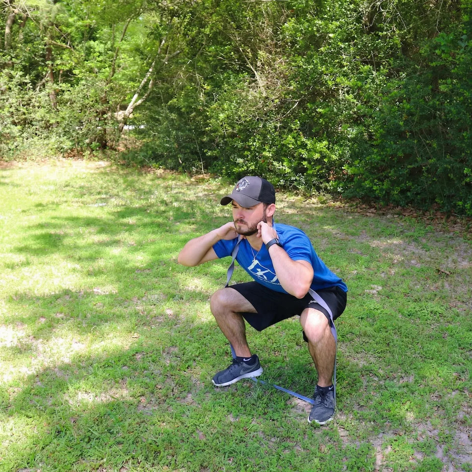 Young athlete performing a front-loaded squat with a Purple TAP® Giant Flat Band anchored under the feet and positioned across the anterior deltoids, creating vertical accommodating resistance for trunk stability and postural organization.