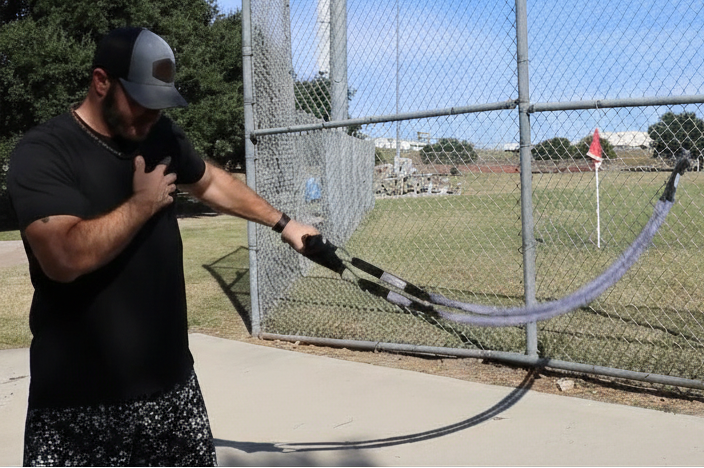 Baseball athlete performing up-down wave exercise with TAP® Sheathed Safety Tubing anchored to chain-link fence.