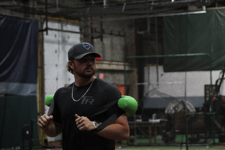 Pitcher from the front performing a pitching delivery with a Bulgarian Water Bag draped over his shoulders, utilizing shifting water resistance to enhance core stability, strength, and dynamic movement control.