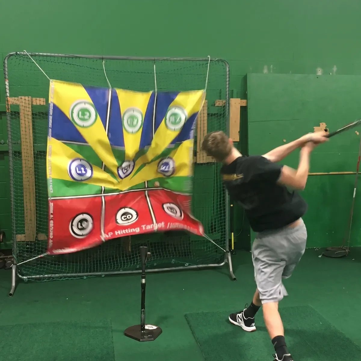 Baseball player taking a powerful swing off a batting tee into the colorful TAP® Hitting Target banner mounted on a net, practicing line drives, fly balls, and ground balls in an indoor training facility.