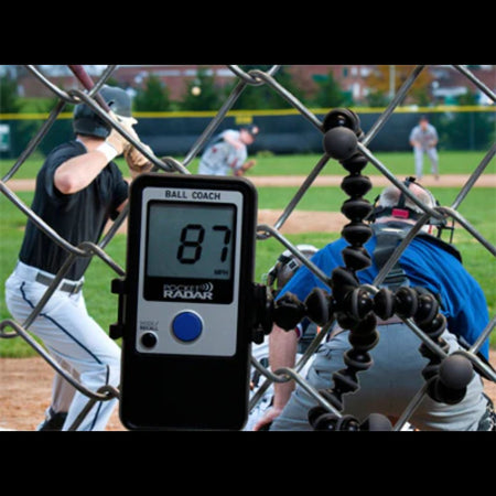 Ball Coach™ radar gun secured to a chain-link fence, actively tracking pitch speeds during a live baseball game—hands-free velocity data for in-game analysis.