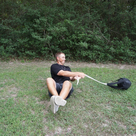 Image depicts a male athlete seated on the grass performing a seated side-to-side slam with light weight medicine ball . He are holding the rope handle as the hammer net and ball is rebounding from the ground. The individual is engaging their core, arms, and shoulders.